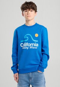 Sininen puuvillaneule, jossa on pyöreä kaula-aukko. Siinä on aalto- ja auringgrafiikka vaaleansinisellä ja oranssilla. "California Making Waves" -teksti valkoisella.