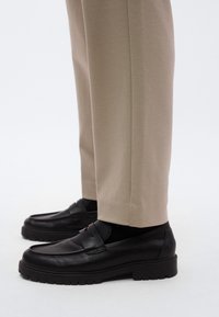 Mocassini neri in pelle con finitura strutturata, caratterizzati da una fascia sul collo del piede e una suola spessa e scolpita, abbinati a pantaloni beige.