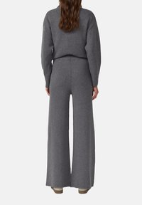 Woman with long brown hair wearing a gray knit sweater and wide-leg pants, shown from the back on a plain background.