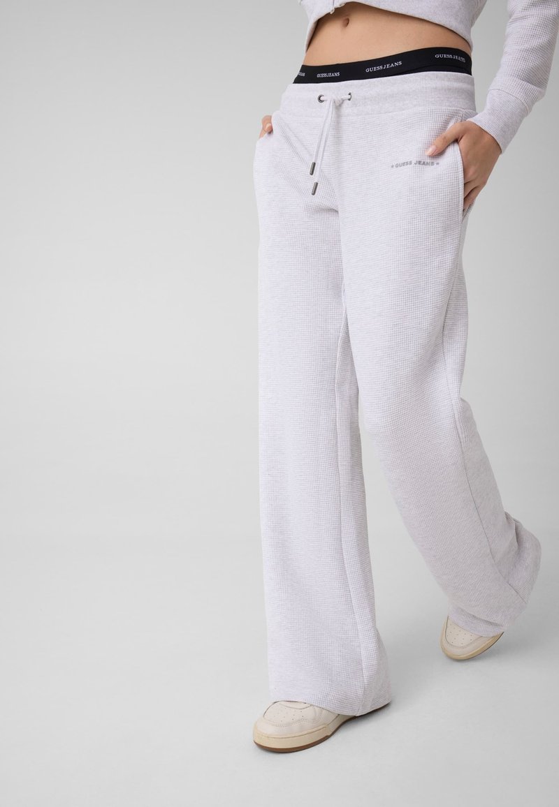 Personne portant un pantalon de survêtement gris clair à cordon, des baskets blanches et un top court, les mains dans les poches, exposant une ceinture noire sur laquelle est écrit "Guess Jeans".