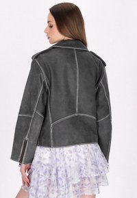 Giacca in suede grigio con un design strutturato, caratterizzata da rever oversize, polsini con zip e cuciture a contrasto. La vista posteriore mostra i dettagli delle cuciture a pannello.
