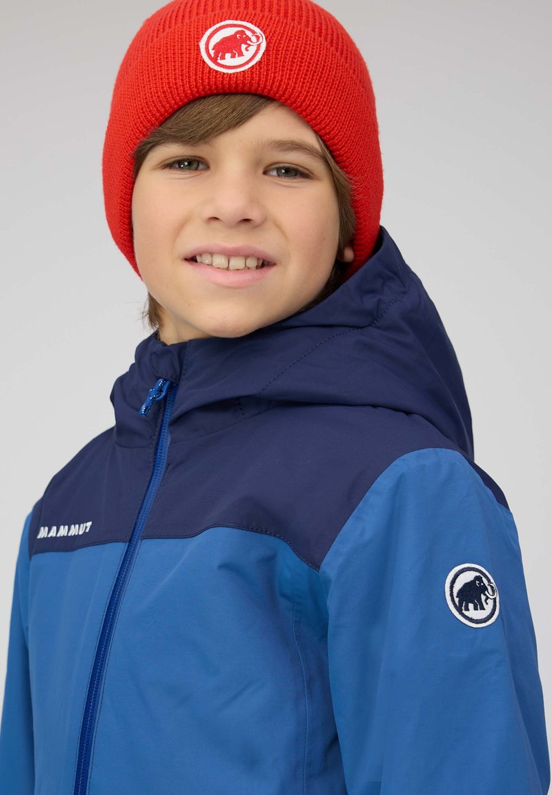 Blaue Jacke mit einem marineblauen Oberteil, ausgestattet mit einem Frontreißverschluss und einem Mammut-Logo. Kombiniert mit einer roten Strickmütze, die ein weißes Logo trägt.