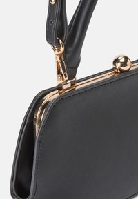 Zwarte leren handtas met een glanzende gouden sluiting, een gestructureerde afwerking en een afgeronde handgreep. Beschikt over een metalen balaccent op de sluiting.