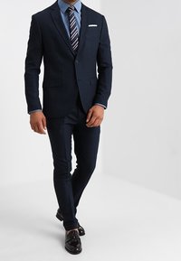 Marineblauw tweedelig pak met een getailleerde pasvorm, gestructureerde stof, bijpassende broek, lichtblauw overhemd, gestreepte stropdas en gepoetste zwarte schoenen.