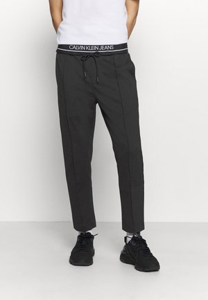 Person trägt schwarze Jogginghosen mit Calvin Klein Jeans-Bund, weiße Socken, schwarze Sneakers und ein weißes Hemd, steht vor schlichtem Hintergrund.