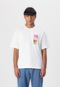 White cotton t-shirt with short sleeves, featuring a pink logo "SR" and a plant graphic with text below, worn with blue jeans.