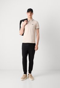 Beige póló fekete kiegészítőkkel, fekete jogger nadrággal és bézs sportcipővel párosítva. A modell egy fekete dzsekit tart az egyik vállán.