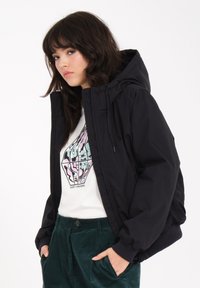 Schwarze Kapuzenjacke mit Reißverschluss, gerippten Bündchen und lockerem Schnitt. Getragen über einem weißen Grafik-T-Shirt und grünen Cordhosen.