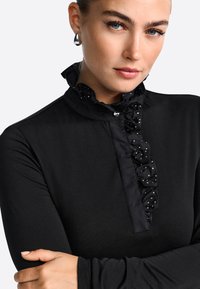 Melnas garas piedurknes blūze ar ruffles apkakli un plāksteri, kurā redzami baltie punktiņi. Mīksta tekstūra un pieguļošs dizains.