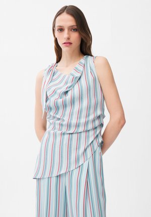 Jeune femme portant un chemisier sans manches bleu clair et un pantalon assorti avec des rayures verticales rouges, blanches et bleues sur un fond uni.