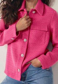 Giacca testurizzata di un brillante colore rosa con un design colletto, caratterizzata da bottoni circolari lucidi e una tasca sul petto. Indossata sopra jeans blu.