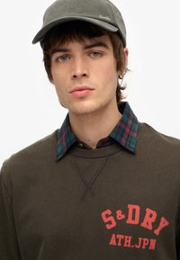Ciemnozielony sweatshirt z czerwonym logo "S&Dry ATH.JPN", nałożony na niebieską i zieloną koszulę w kratę z kołnierzykiem, w zestawie z szarym czapką.