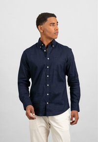 Marineblå skjorte med knapper og lange ermer, liten brystlomme og hvite knapper. Stoffet ser glatt og lett ut.