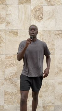 Camiseta gris de manga corta combinada con pantalones cortos negros; persona sosteniendo un micrófono frente a un fondo de pared de piedra texturizada.