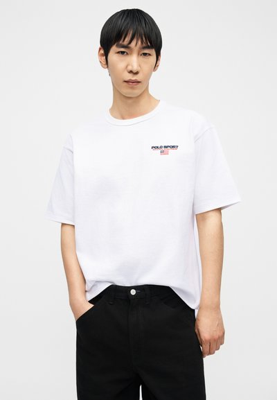 Άνδρας με μαύρα μαλλιά, ντυμένος με λευκή μπλούζα Polo Sport και μαύρο παντελόνι, στέκεται με το ένα χέρι στην τσέπη μπροστά σε απλό φόντο.