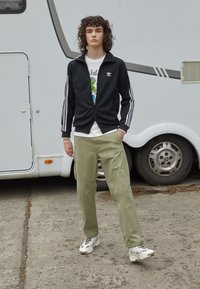 Svart Adidas treningsjakke med hvite striper, hvit grafisk T-skjorte, olivenfargede cargo-bukser og beige sneakers. Utendørs setting.