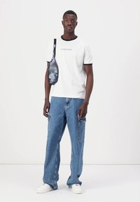 Camiseta blanca con detalles en negro, pantalones de cargo de mezclilla azul y un bolso bandolera de patrón negro y gris, llevados con zapatillas blancas.