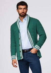 Cárdigan verde de punto trenzado con acentos de franjas blancas en el cuello chal y los puños, combinado con una camisa de rayas azul clara y jeans oscuros.