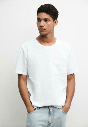 Jeune homme aux cheveux bouclés portant un t-shirt blanc avec poche et un jean bleu clair, debout, les mains dans les poches, regardant vers sa gauche.