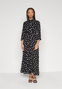 Robe maxi noire à pois avec un col et une fermeture à boutons sur le devant. Présente des manches longues et une silhouette fluide.