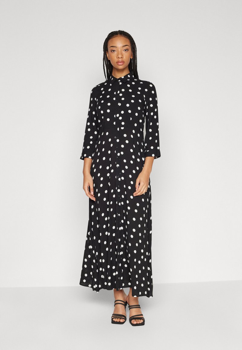 Robe maxi noire à pois avec un col et une fermeture à boutons sur le devant. Présente des manches longues et une silhouette fluide.