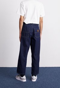 Ciemnoniebieskie dżinsy o luźnym kroju, z widocznymi tylnymi kieszeniami i beżową metką. Połączone z prostą białą koszulką i szarymi trampkami.