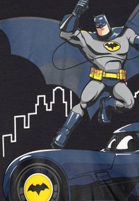 Grafica di Batman su tessuto scuro; presenta tonalità di blu e grigio, skyline della città e simbolo del pipistrello giallo. Include dettagli sul costume e sugli elementi del veicolo.