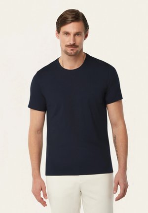 Mann steht und trägt ein einfaches, marineblaues, kurzärmliges T-Shirt und weiße Hosen, mit neutralem Gesichtsausdruck und Tätowierung auf seinem rechten Unterarm.