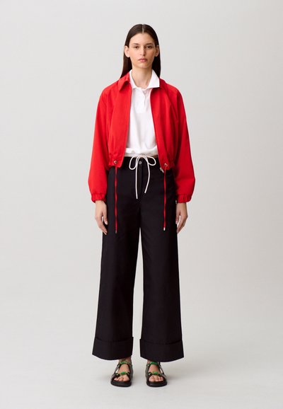 Giacca corta rossa con polsini elasticizzati, polo bianca, pantaloni neri a gamba larga con coulisse e sandali neri con dettagli verdi.