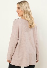 Vaaleanpunainen neulepaita, joka on valmistettu pehmeästä kankaasta. Paidassa on väljä malli, pitkät hihat ja koristeelliset helmivälikkeet olkasaumojen kohdalla.