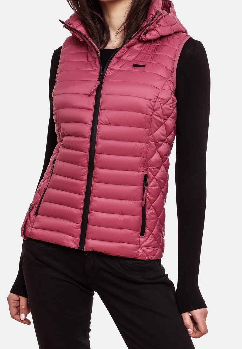 Femme portant une veste sans manches matelassée rose avec capuche, sur une chemise noire à manches longues et un pantalon noir.