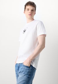 Camiseta blanca de algodón con un gráfico negro en el pecho, mangas cortas y un escote redondo, combinada con jeans de mezclilla azul claro.