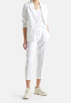 Persona con blazer blanco, top blanco, pantalones blancos recortados, zapatillas gruesas beige y un collar dorado con colgantes de colores.