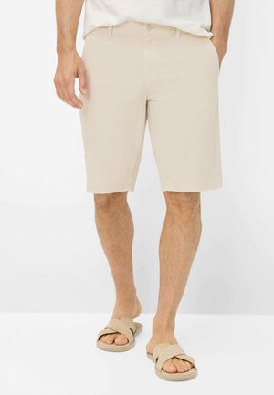 Mand iført beige knælange shorts, hvid skjorte og matchende beige åbne sandaler, stående mod en ensfarvet hvid baggrund.