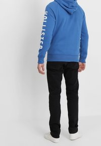 Blå huvtröja med "Hollister" tryckt i vitt på vänster ärm, matchad med svarta byxor och vita sneakers.