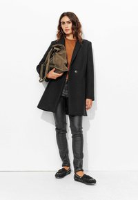 Manteau en laine noir, pull marron, pantalon en cuir noir et chaussures noires ornées. La mannequin tient un sac vert olive. Fond blanc.