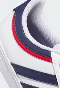 Białe sneakersy z granatowymi i czerwonymi akcentami, wykonane z materiału syntetycznego, o teksturowanej powierzchni i warstwowych paskach po bokach.