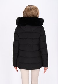 Jachetă puffer neagră cu guler din blană artificială, având un design matlasat, glugă și mâneci lungi, purtată peste pantaloni bej.