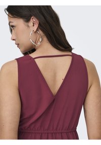 Robe sans manches bordeaux avec un design de dos en V, taille froncée et tissu texturé ; accompagnée de grandes boucles d'oreilles en argent.