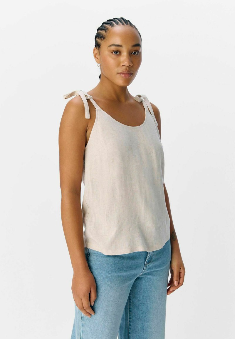 Haut sans manches beige clair avec un col rond et des bretelles à nouer, fabriqué à partir d'un tissu doux et texturé. Associé à un jean taille haute bleu.