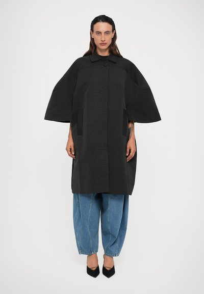 Donna che indossa un cappotto nero oversize con texture e maniche larghe, jeans chiari larghi e décolleté neri a punta, in piedi contro uno sfondo bianco.