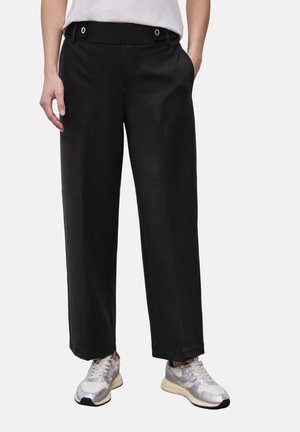 Person iført sorte bukser med lige ben, knappede bæltestropper og sølvfarvede sneakers, stående med venstre hånd i lommen.