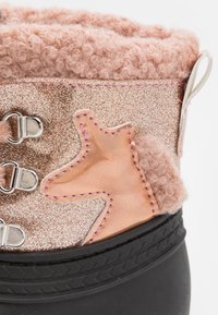La bota presenta una parte superior metálica en color rosa dorado con un puño de terciopelo rosa, costuras decorativas y una suela negra texturizada. Acabada con una forma rosa.