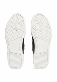 Black sneakers with a textured upper and white rubber sole featuring circular patterns and a herringbone tread. Subtle logo detail on the sole.