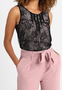 Černý krajkový top bez rukávů s průsvitnou strukturou, zdobený sklady a květinovými vzory, nošený s růžovými kalhotami s vysokým pasem a uvázaným opaskem.