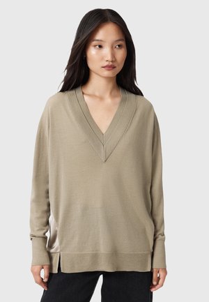 Jeune femme aux longs cheveux bruns, portant un pull beige à col en V ample et un pantalon noir, se tenant devant un fond gris clair.