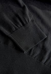 Sort sweater med ribbede manchetter og tekstureret stof. Nærbillede fremhæver den glatte strik og let samlet ærmedesign.