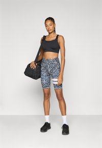 Svart sportbh, mønstrede sykkelshorts i blått og hvitt, svarte sko, holder en svart veske, står mot en nøytral bakgrunn.