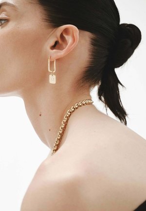 Perfil de una mujer con cabello oscuro recogido en un moño, que lleva un collar de cadena dorada y un pendiente de aro rectangular dorado con un pequeño charm cuadrado.