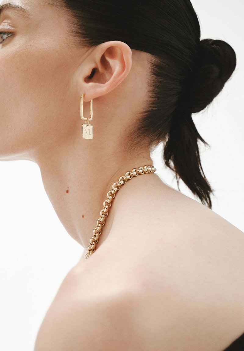 Perfil de una mujer con cabello oscuro recogido en un moño, que lleva un collar de cadena dorada y un pendiente de aro rectangular dorado con un pequeño charm cuadrado.